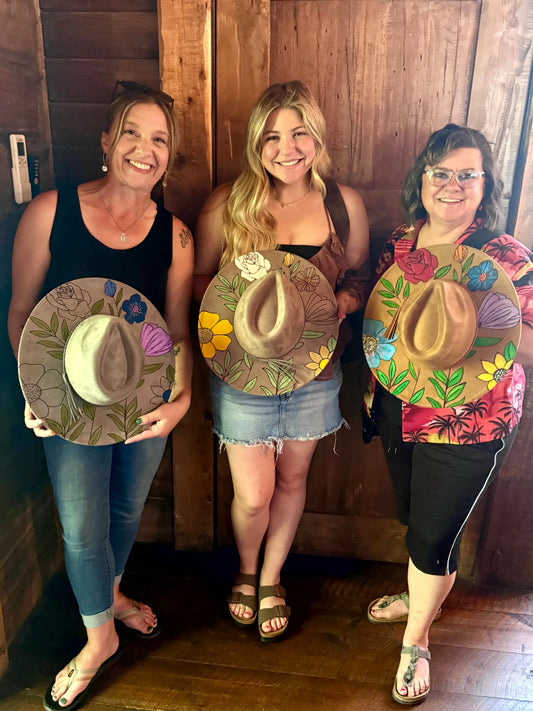 Three women proudly display their custom-painted wide-brim hats featuring laser-burned floral designs.