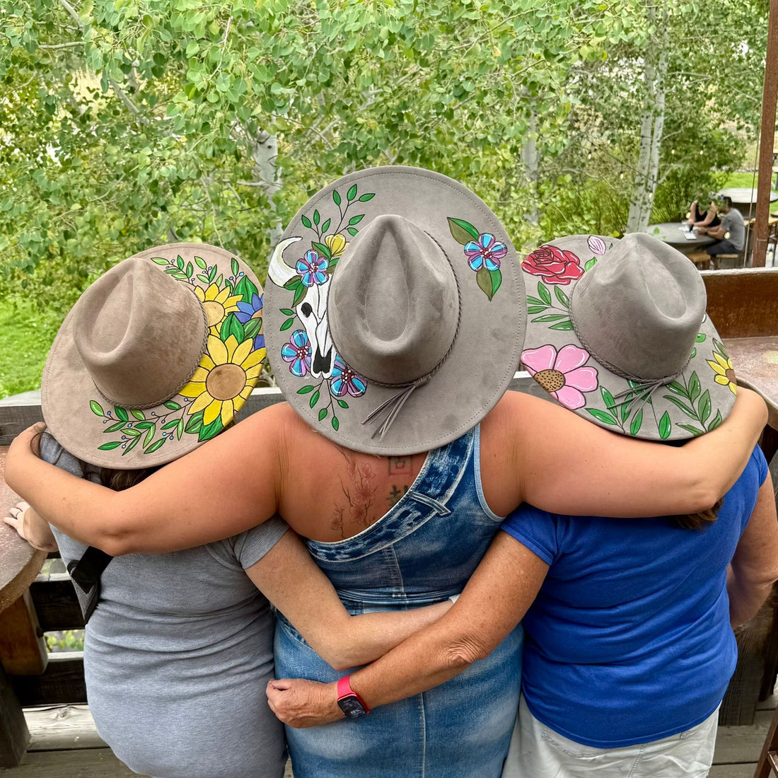 A group of friends wearing custom-designed wide-brim hats with sunflowers, steer skulls, and wildflower motifs